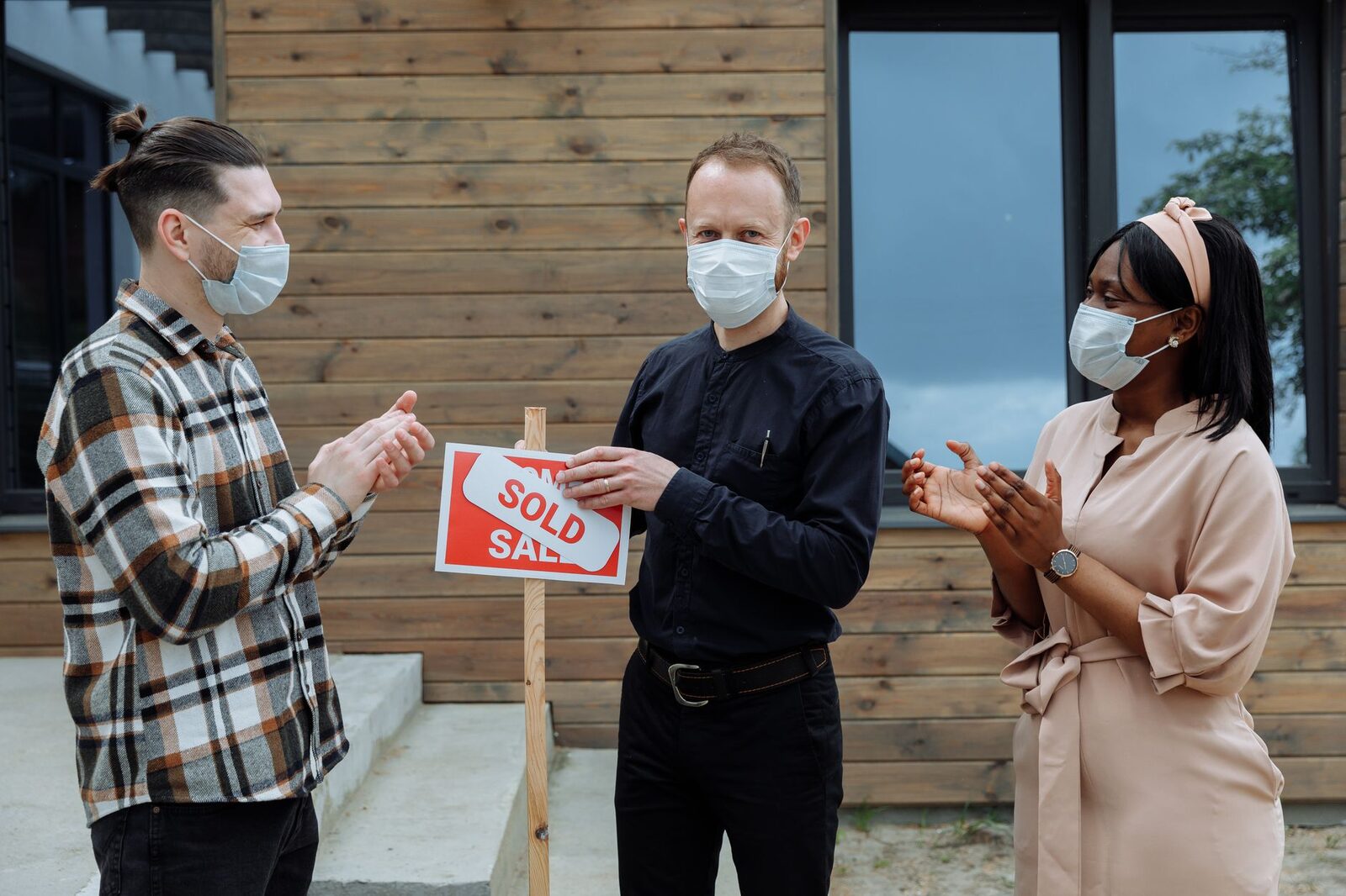 Real estate agent and clients celebrate sale with masks and a sold sign outdoors.
